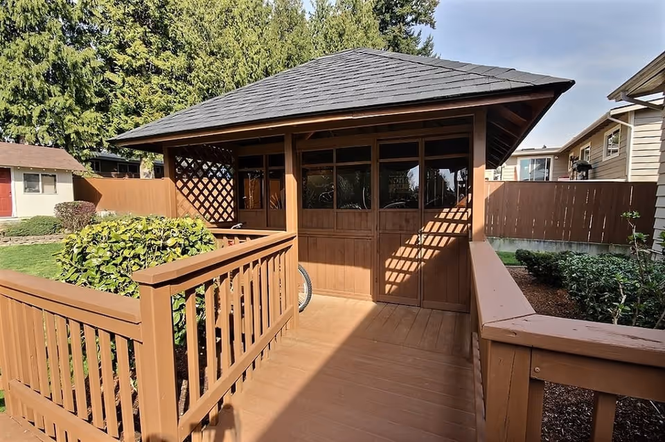 A wooden gazebo with a shingled roof situated in a garden area. The gazebo has wooden railings and lattice panels on the sides, surrounded by green bushes and trees. Nearby houses and a wooden fence are visible in the background under a partly cloudy sky.
