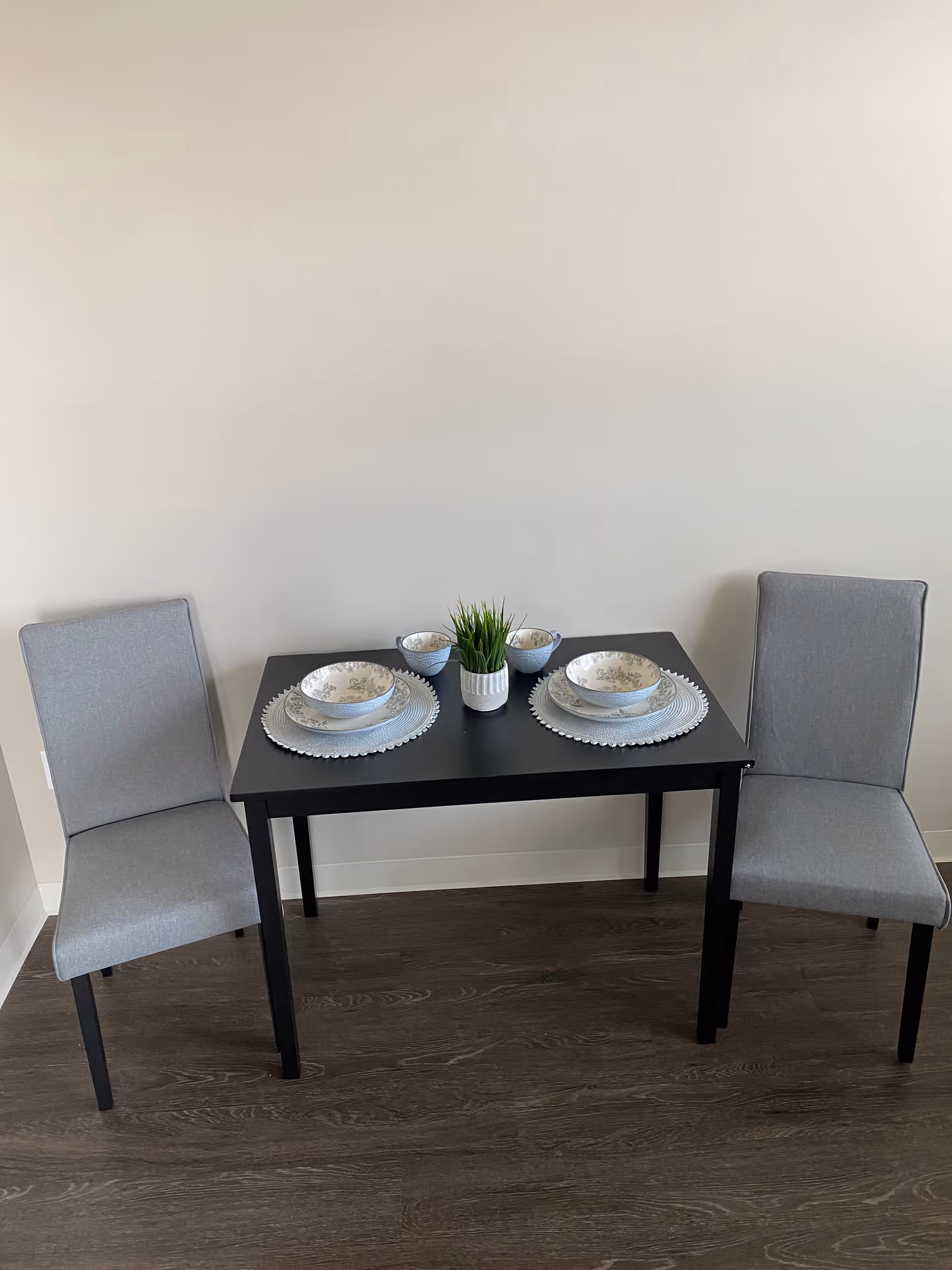A small black dining table set for two with two grey upholstered chairs. The table has two place settings with white and grey patterned bowls and plates, two small matching cups, and a small potted green plant in the center. The floor is dark wood and the wall behind is plain white.