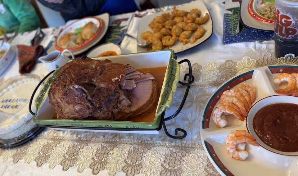 A dining table set with various dishes including a large piece of cooked ham in a green and white serving dish, a plate of cooked shrimp with cocktail sauce, a plate of fried shrimp, and a plate with crescent rolls. The table is covered with a white lace tablecloth and there are people seated around the table.