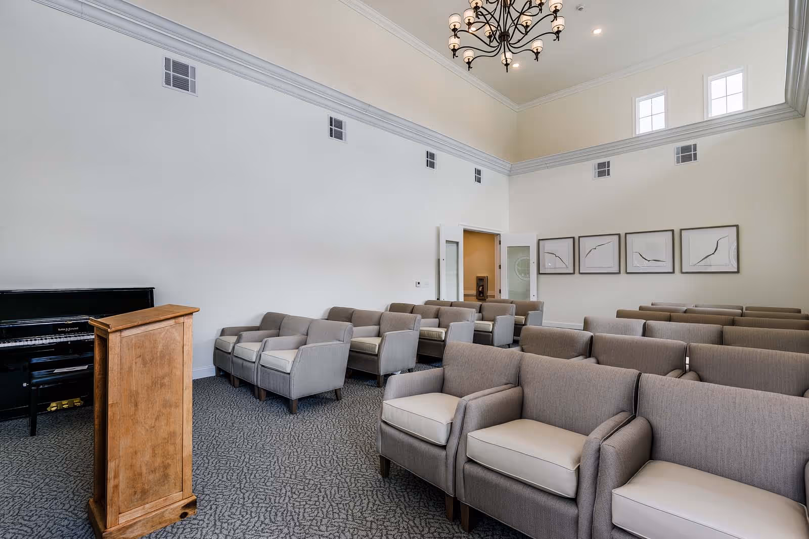 A room with multiple rows of gray cushioned armchairs arranged facing a wooden podium. There is a black upright piano against the left wall. The room has high ceilings with a decorative chandelier and several small windows near the ceiling. Four framed abstract artworks hang on the far wall.