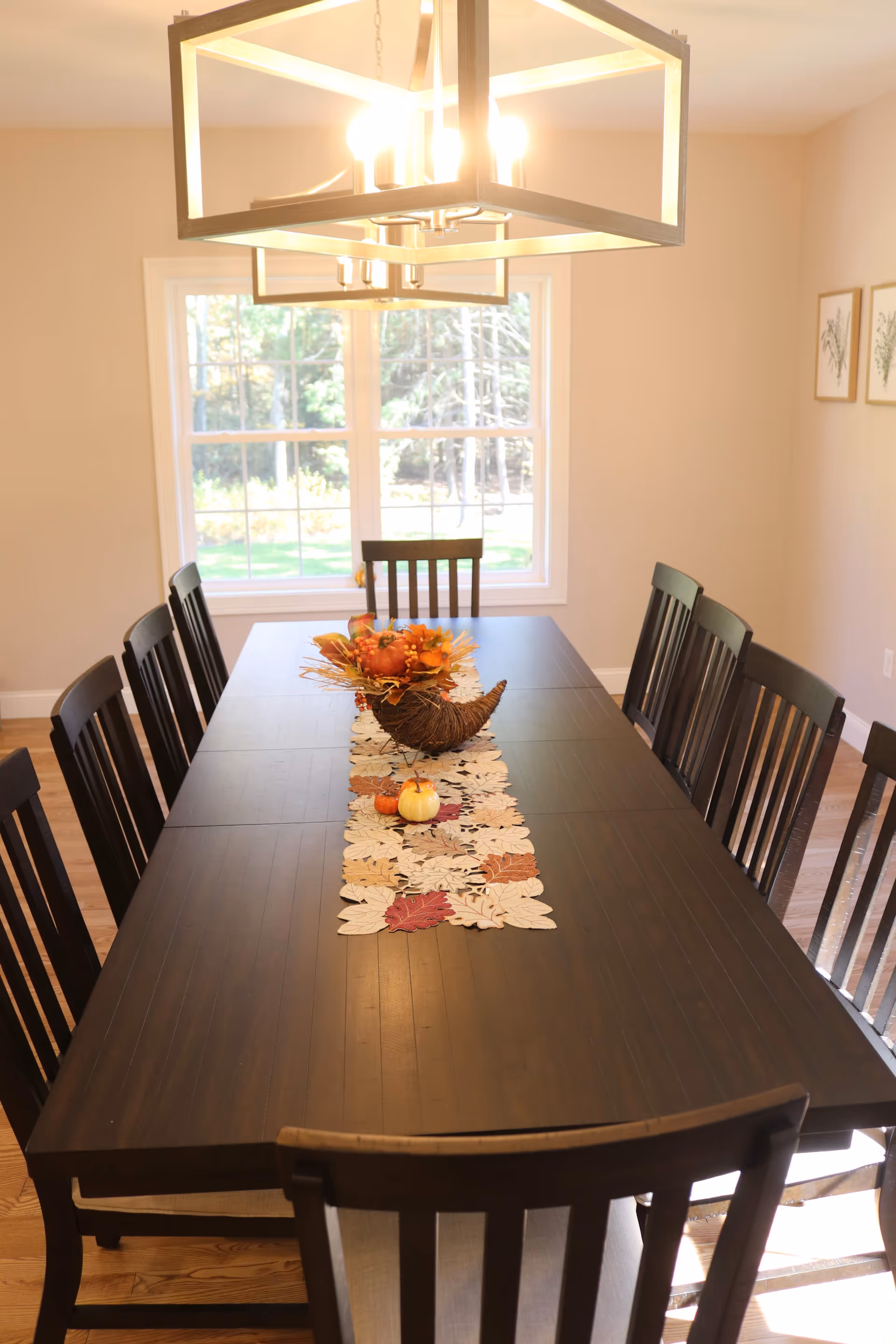 Long dark wood dining table with chairs, a fall centerpiece, and a large window under a modern chandelier.
