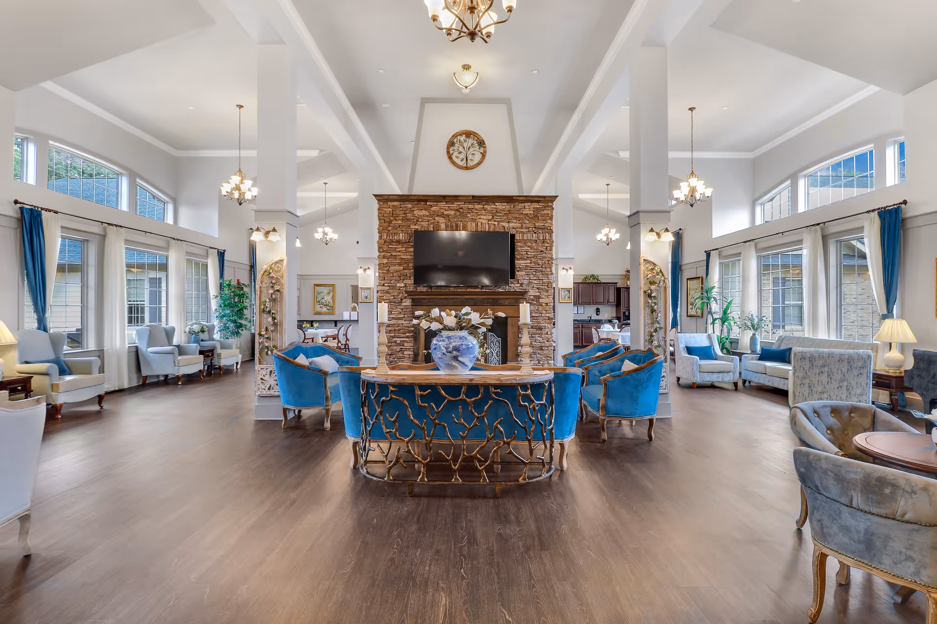 Spacious and bright senior living common area with high ceilings, large windows with white and blue curtains, multiple seating arrangements including blue and gray armchairs and sofas, a stone fireplace with a mounted TV above it, decorative plants, and elegant chandeliers hanging from the ceiling.