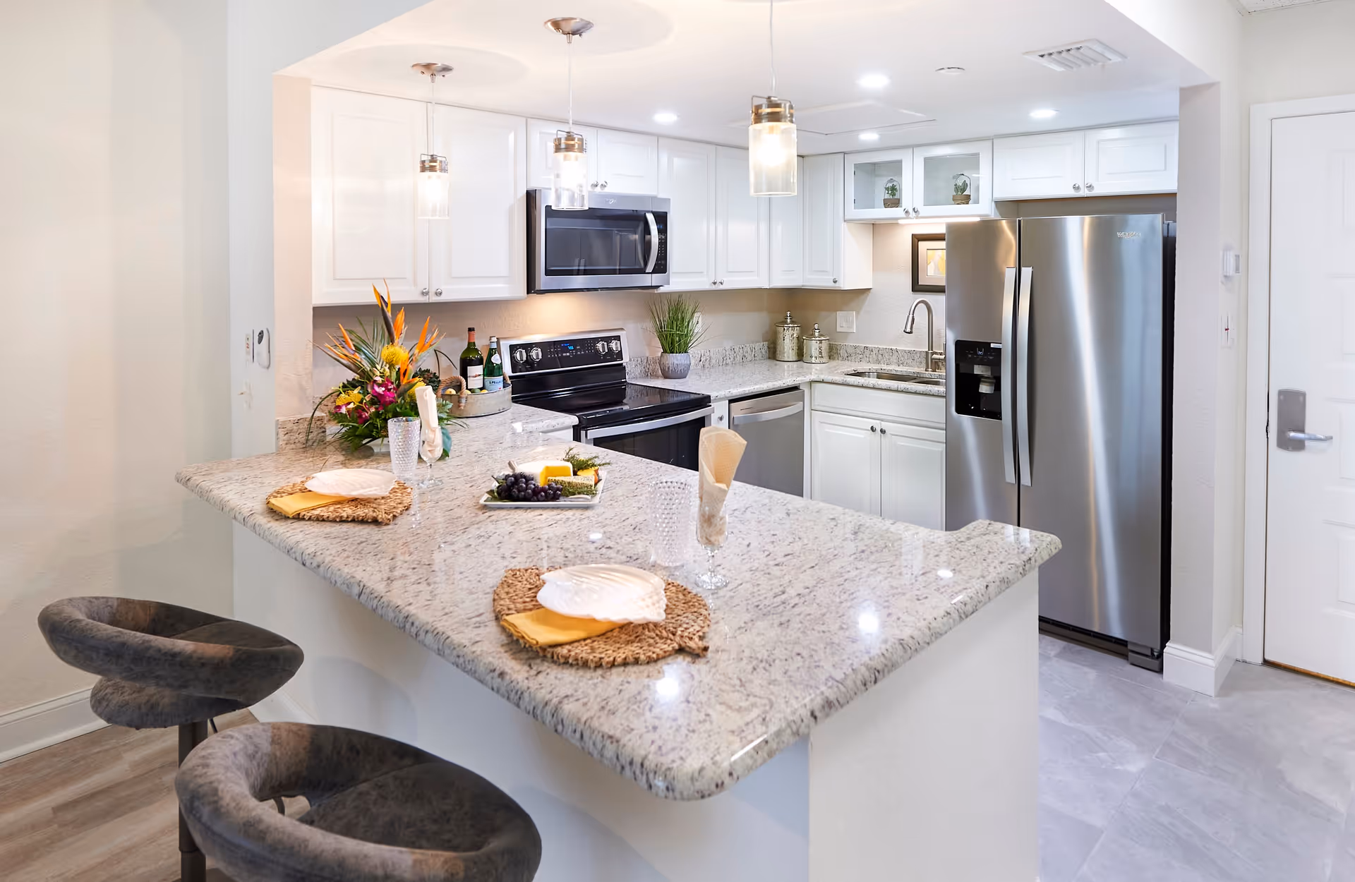 Modern kitchen with white cabinets, granite countertops, stainless steel appliances including a refrigerator, oven, microwave, and dishwasher. The kitchen island has two gray cushioned bar stools and is set with plates, glasses, and napkins. There are three pendant lights hanging above the island and a floral arrangement on the counter.