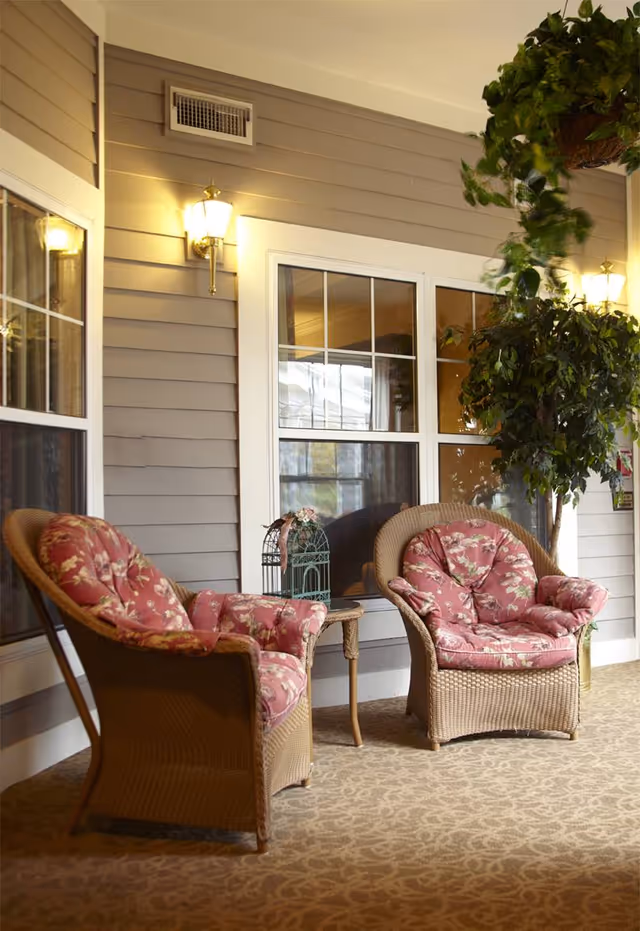 A cozy indoor seating area with two wicker armchairs featuring floral pink cushions, a small wooden side table with a decorative birdcage, wall-mounted lantern-style lights, large windows, and a hanging green plant.