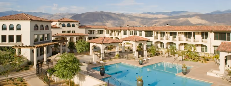 Outdoor view of a senior living facility named Segovia featuring a large swimming pool surrounded by patio furniture, potted plants, and a multi-story building with arched windows and terracotta roof tiles, set against a backdrop of mountains under a partly cloudy sky.