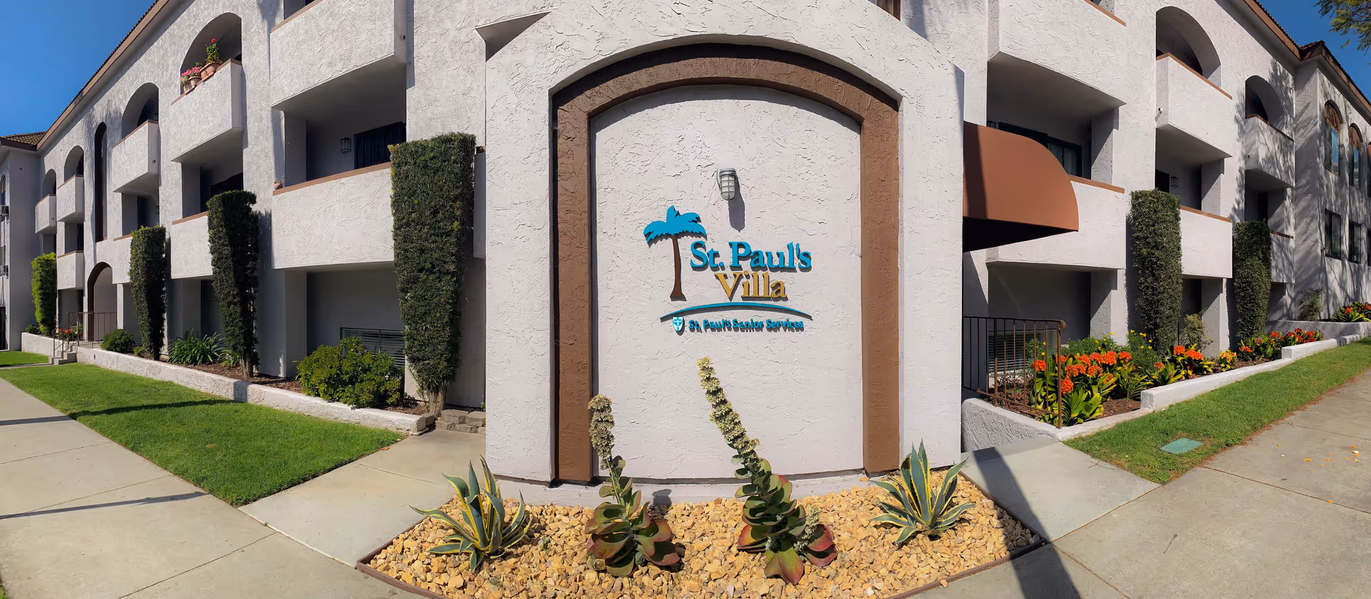 Exterior view of a two-story residential building with white stucco walls and brown trim. The building has balconies with arches and is surrounded by neatly trimmed bushes, green grass, and flower beds. A sign on the wall reads 'St. Paul's Villa' with a palm tree graphic above it.