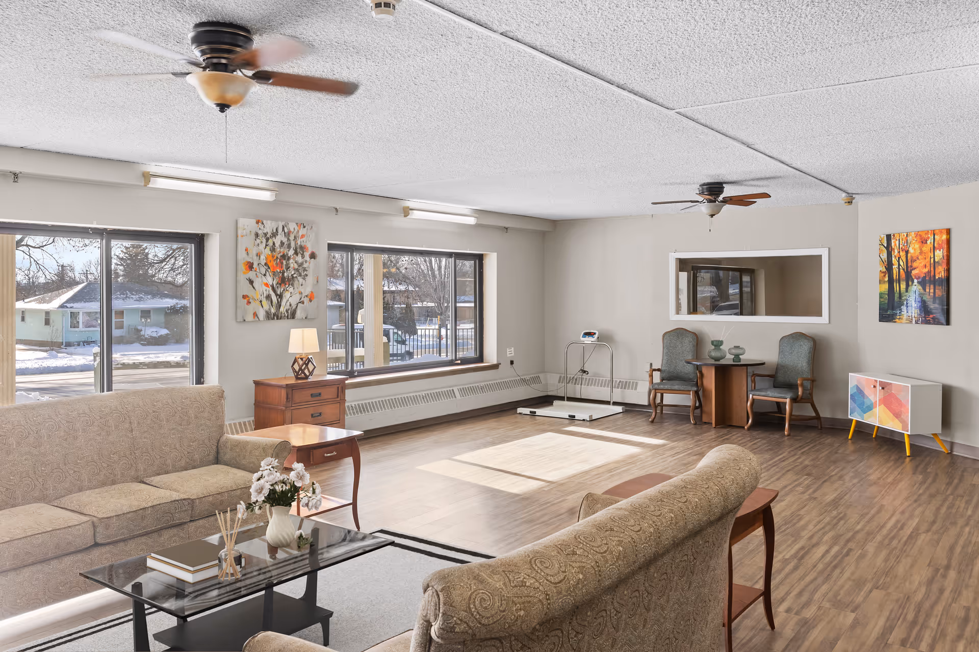A spacious living room with two beige patterned sofas facing each other, a glass coffee table with books and a flower vase, wooden side tables, and large windows letting in natural light. The room has wooden flooring, two ceiling fans, wall art, and a small table with two chairs in the corner.