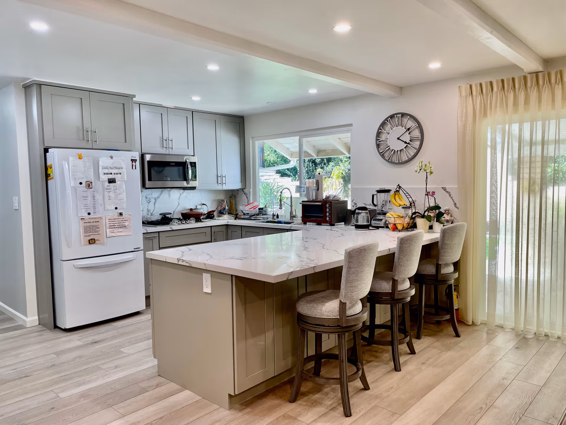 A modern kitchen with light gray cabinets, a white refrigerator with papers attached, a built-in microwave, and a large marble countertop island with three cushioned bar stools. There is a window above the sink showing greenery outside, a wall clock, and sheer curtains covering a sliding glass door.
