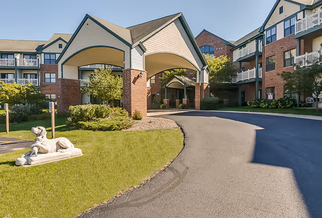 Exterior view of Meadowmere & Mitchell Manor Oak Creek showing a driveway leading to a covered entrance between two multi-story brick and siding buildings with balconies. There is a lawn area with a statue of a dog lying down near the driveway.