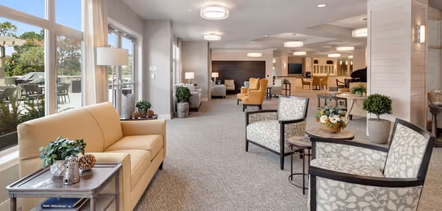A spacious and well-lit senior living facility common area with large windows on the left letting in natural light. The room features a beige sofa, patterned armchairs, small tables with plants and decorative items, and additional seating further back. The decor is modern and inviting with neutral tones and soft lighting.