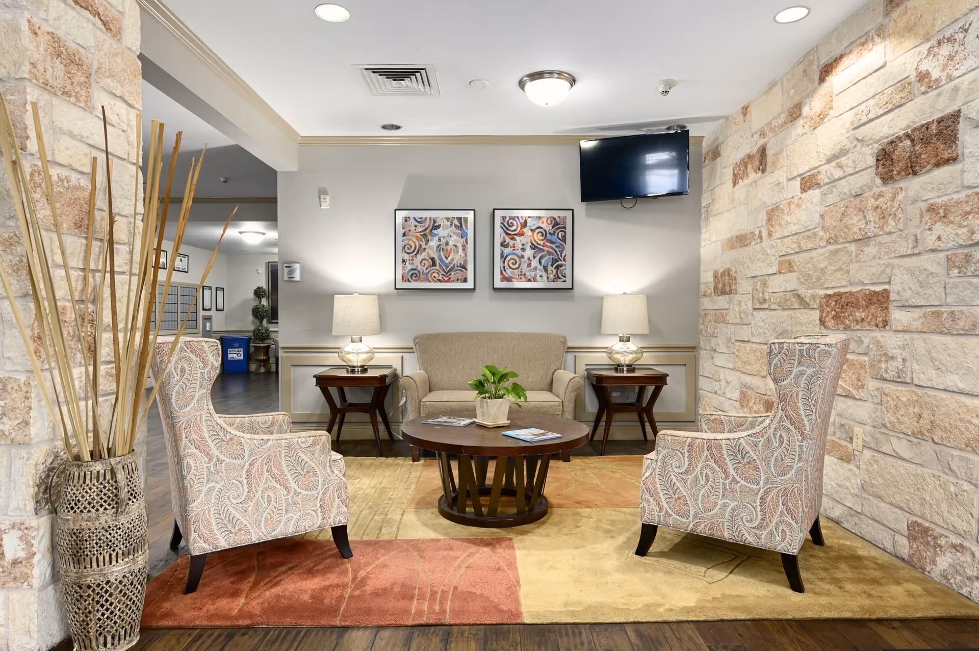 A cozy seating area in a senior living facility featuring two patterned armchairs and a beige loveseat arranged around a round wooden coffee table with a small potted plant. The space has a stone accent wall, two table lamps on wooden side tables, two framed abstract artworks on the wall, and a wall-mounted TV above the loveseat. The floor has a multicolored area rug and wooden flooring.