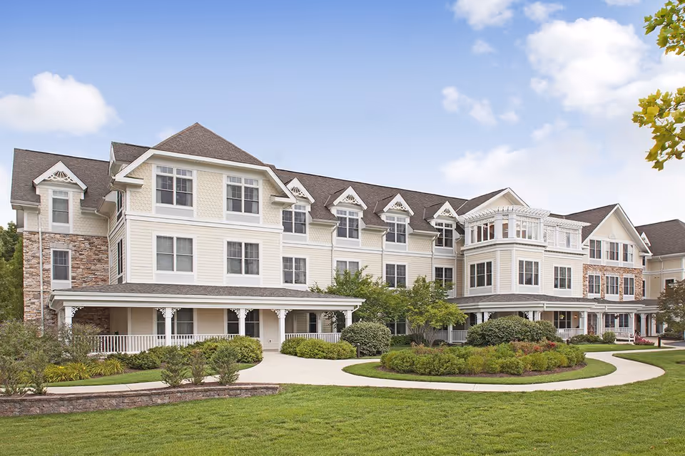 Exterior view of a large, multi-story senior living facility building with beige siding, stone accents, multiple windows, and a covered porch. The building is surrounded by well-maintained green lawns, shrubs, and trees under a partly cloudy sky.