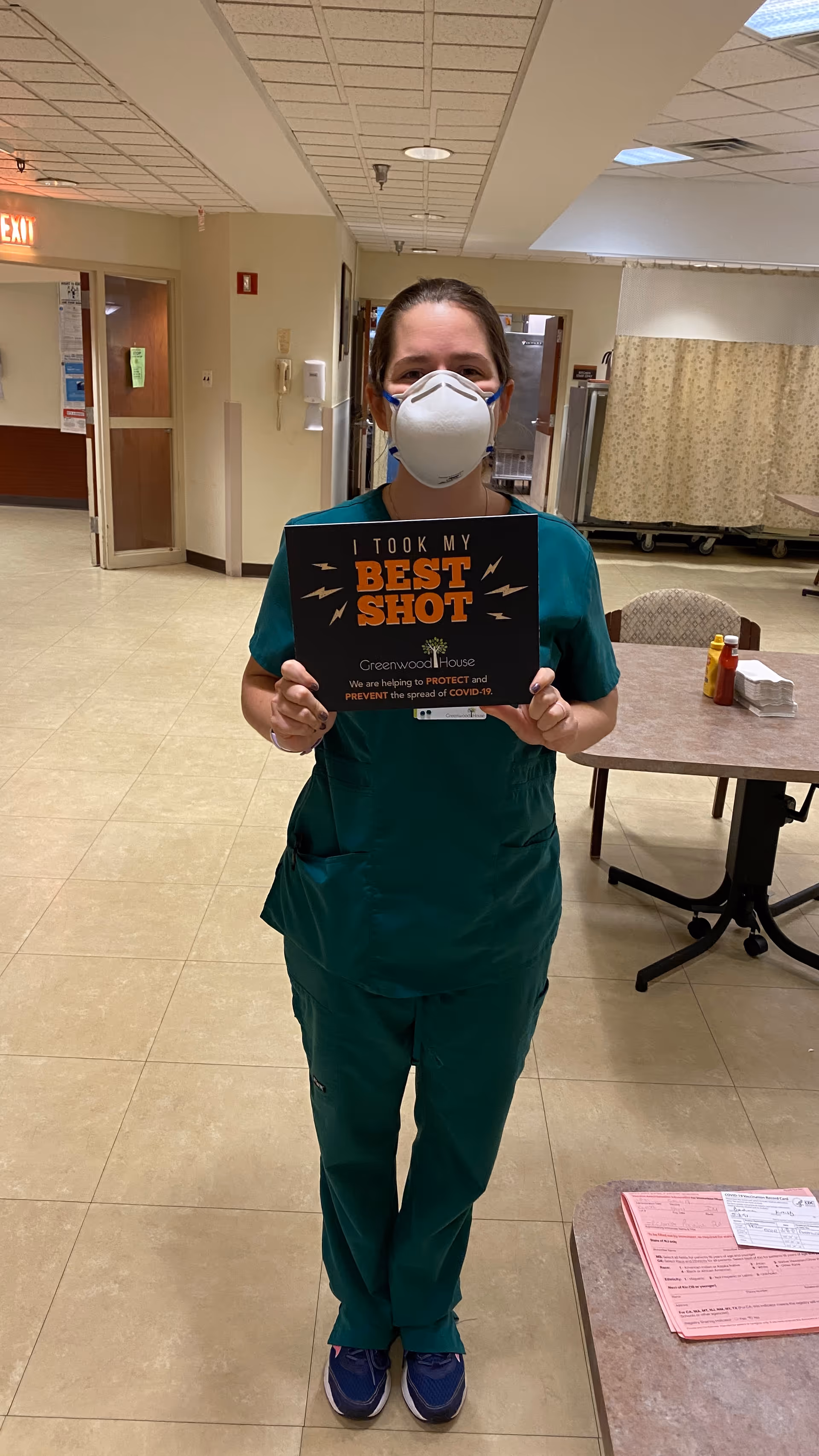 A healthcare worker wearing green scrubs and a white face mask stands in a hallway holding a sign that reads 'I TOOK MY BEST SHOT Greenwood House We are helping to PROTECT and PREVENT the spread of COVID-19.' The background shows a table with condiments and papers, chairs, and a curtain divider.