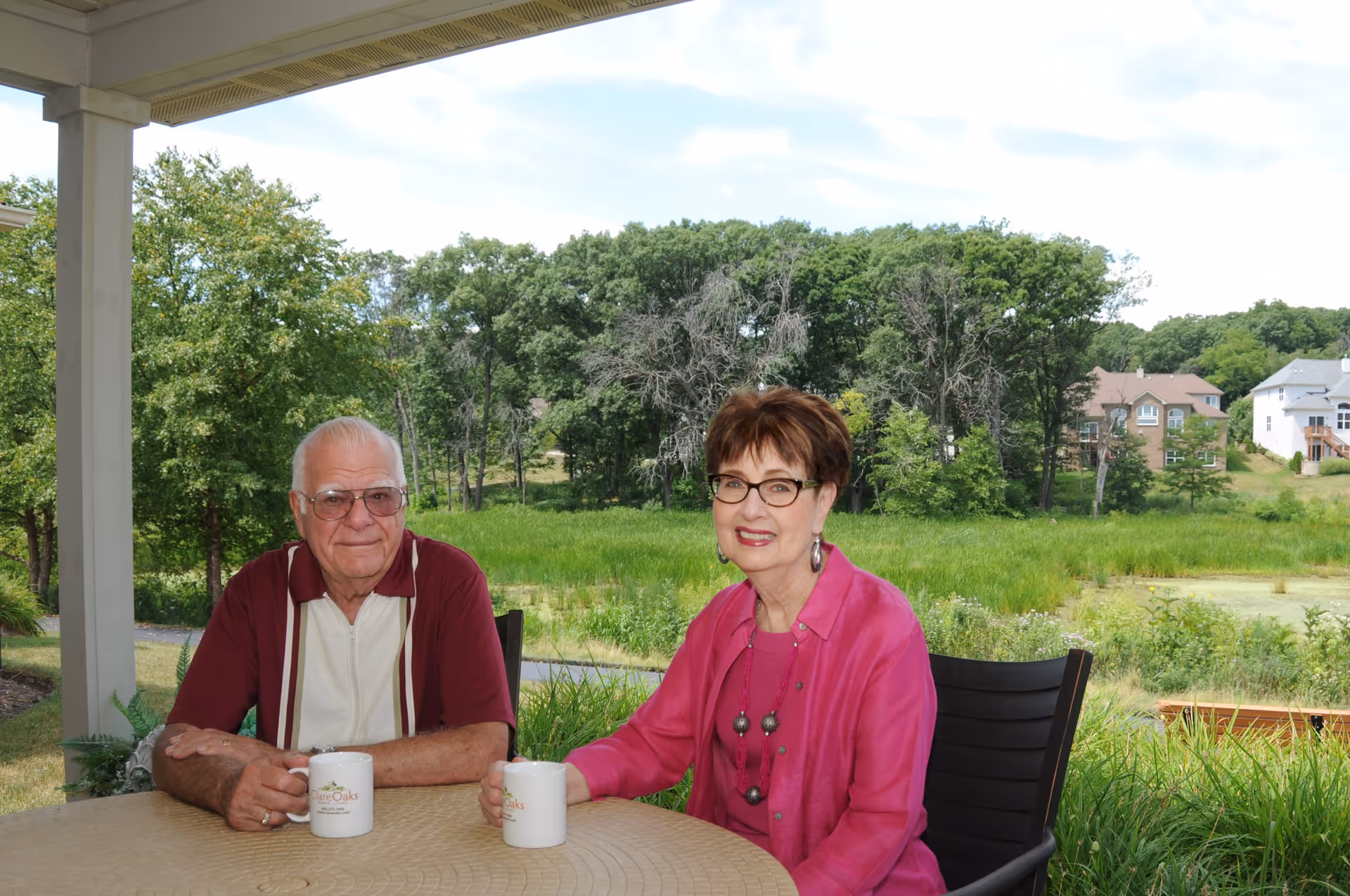 An elderly man and woman sitting at a round outdoor table under a covered patio. Both are holding white mugs with 'The Oaks' logo. Behind them is a green grassy area with trees and houses in the background under a partly cloudy sky.