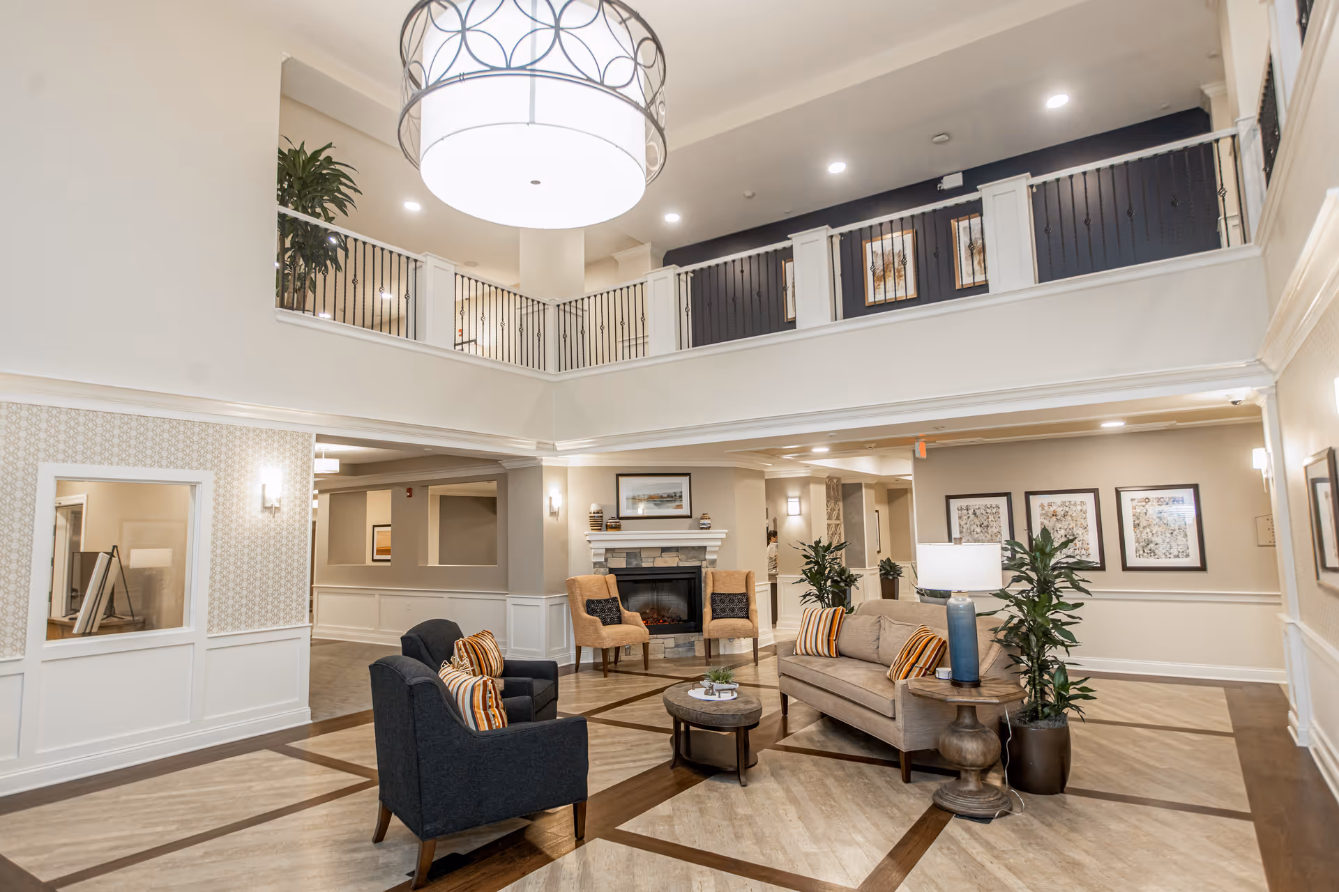 A spacious and well-lit senior living facility common area with a high ceiling and a large hanging light fixture. The room features a seating arrangement with a beige sofa, two dark armchairs, and two tan chairs around a round coffee table. There is a fireplace with decorative items on the mantel, potted plants, framed artwork on the walls, and a balcony railing overlooking the area.