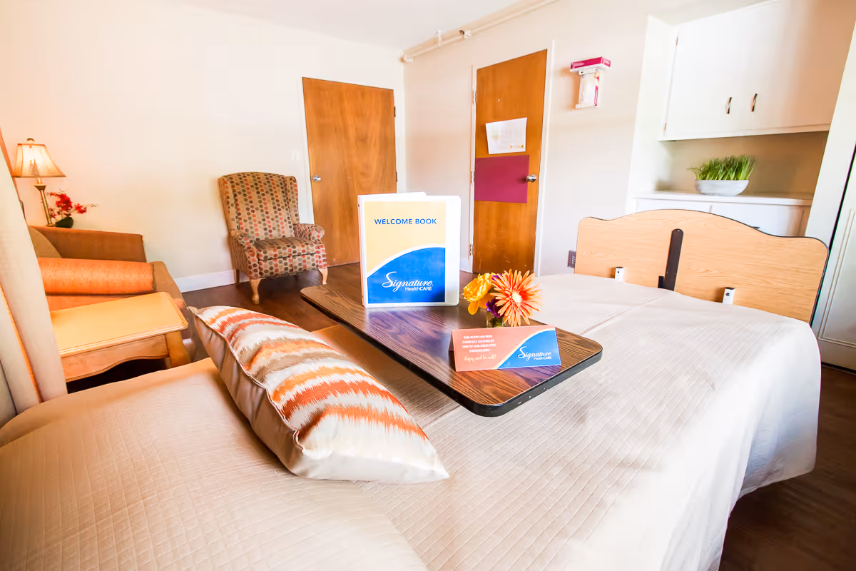A well-lit room in a senior care facility featuring a hospital-style bed with a white quilt and a patterned pillow. On the bed is a wooden overbed table holding a welcome book and a small card with the Signature HealthCARE logo, along with a small flower arrangement. In the background, there is a patterned armchair, a wooden door, a lamp with a red flower decoration, and white cabinetry with a bowl of green plants.