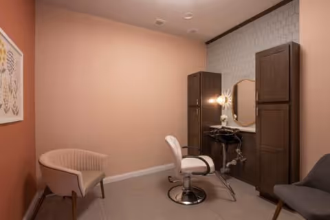 A small room with muted pink walls featuring a salon-style chair in front of a vanity with a mirror and two tall wooden cabinets on either side. There are two additional chairs, one beige and one dark gray, positioned against the walls. The floor is tiled in a neutral color.