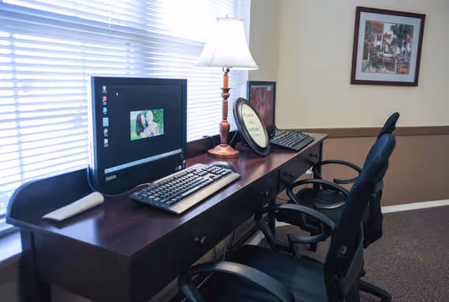 A workspace area with a long wooden desk holding two computer monitors, keyboards, a table lamp, and a framed picture. Two black office chairs are positioned in front of the desk. A window with blinds is behind the desk, and a framed artwork hangs on the beige wall.