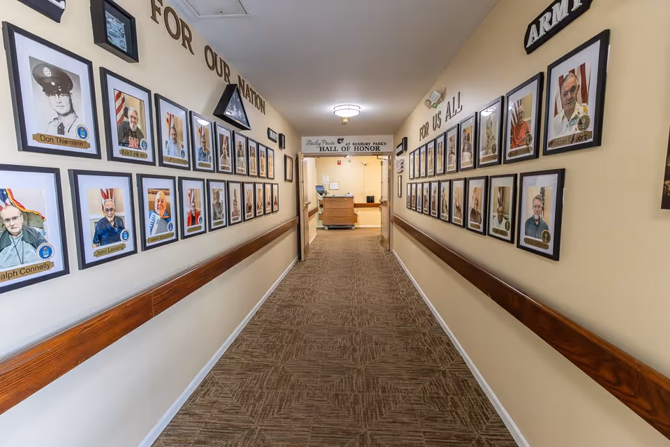 A hallway in Bailey Pointe at Roxbury Park featuring a 'Hall of Honor' display with framed photographs of veterans on both walls. The walls have inscriptions reading 'FOR OUR NATION' and 'FOR US ALL' above the photos. The hallway has beige walls, a patterned carpet, and wooden handrails on both sides.