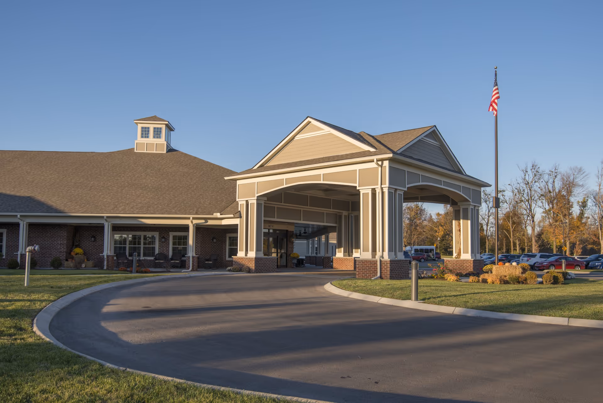 Exterior view of The Lantern at Morning Pointe Alzheimer's Center of Excellence in Danville, showing the building entrance with a covered driveway, an American flag on a flagpole, and a parking lot with cars in the background under a clear blue sky.