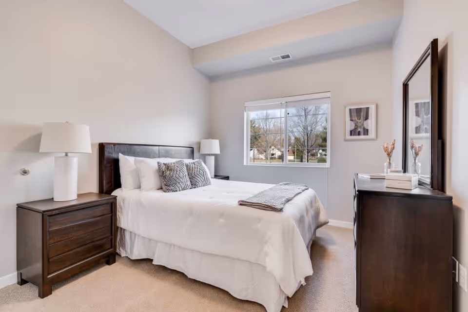 Well-lit bedroom with a bed, two bedside tables with lamps, a dresser with mirror, and a window.