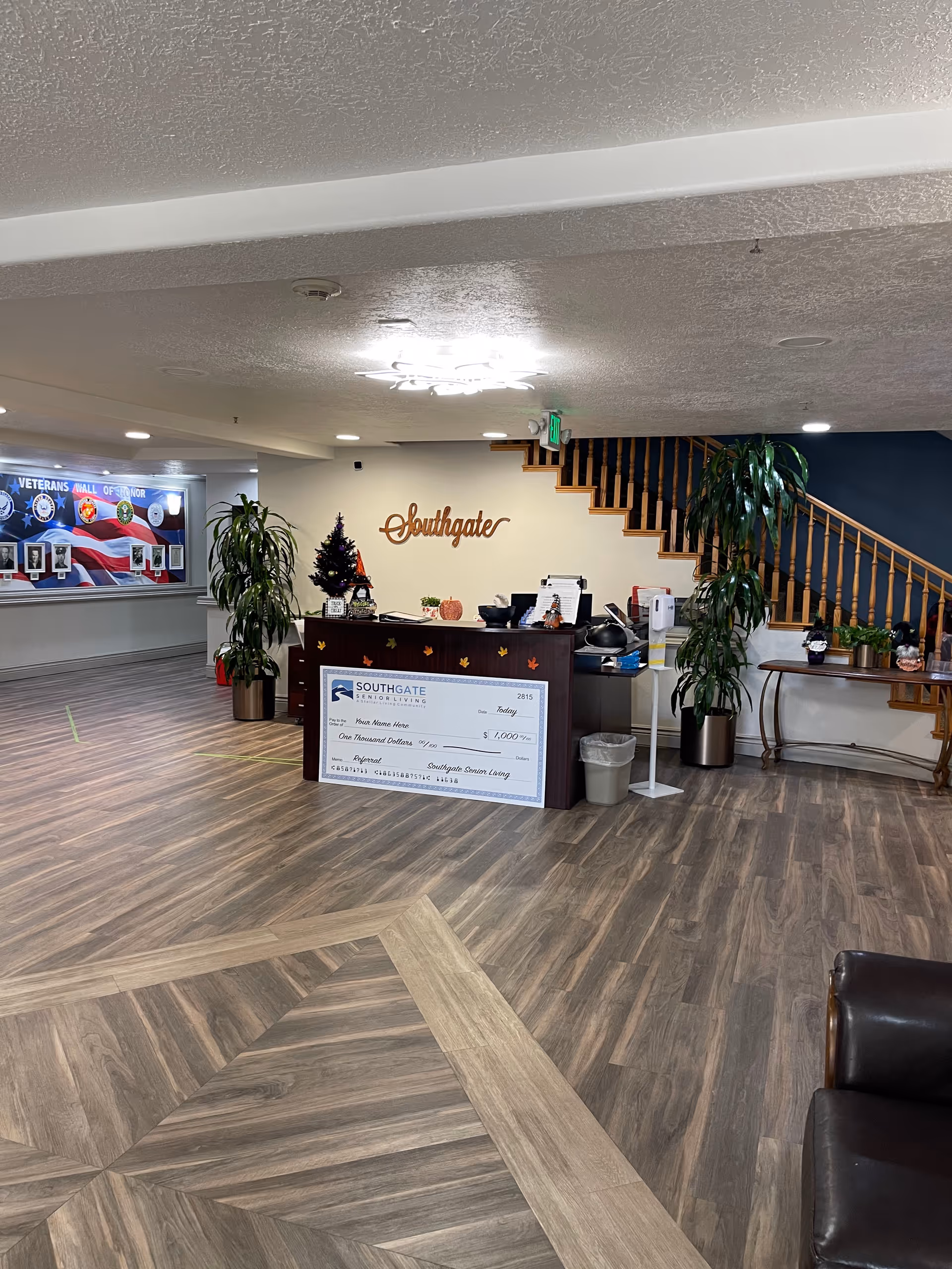 Interior view of the Southgate Senior Living facility lobby area featuring a reception desk with a large decorative check, plants, a small decorated Christmas tree, and a staircase with wooden railings. The wall behind the desk has the word 'Southgate' mounted on it, and there is a Veterans Wall of Honor display on the left side of the image.