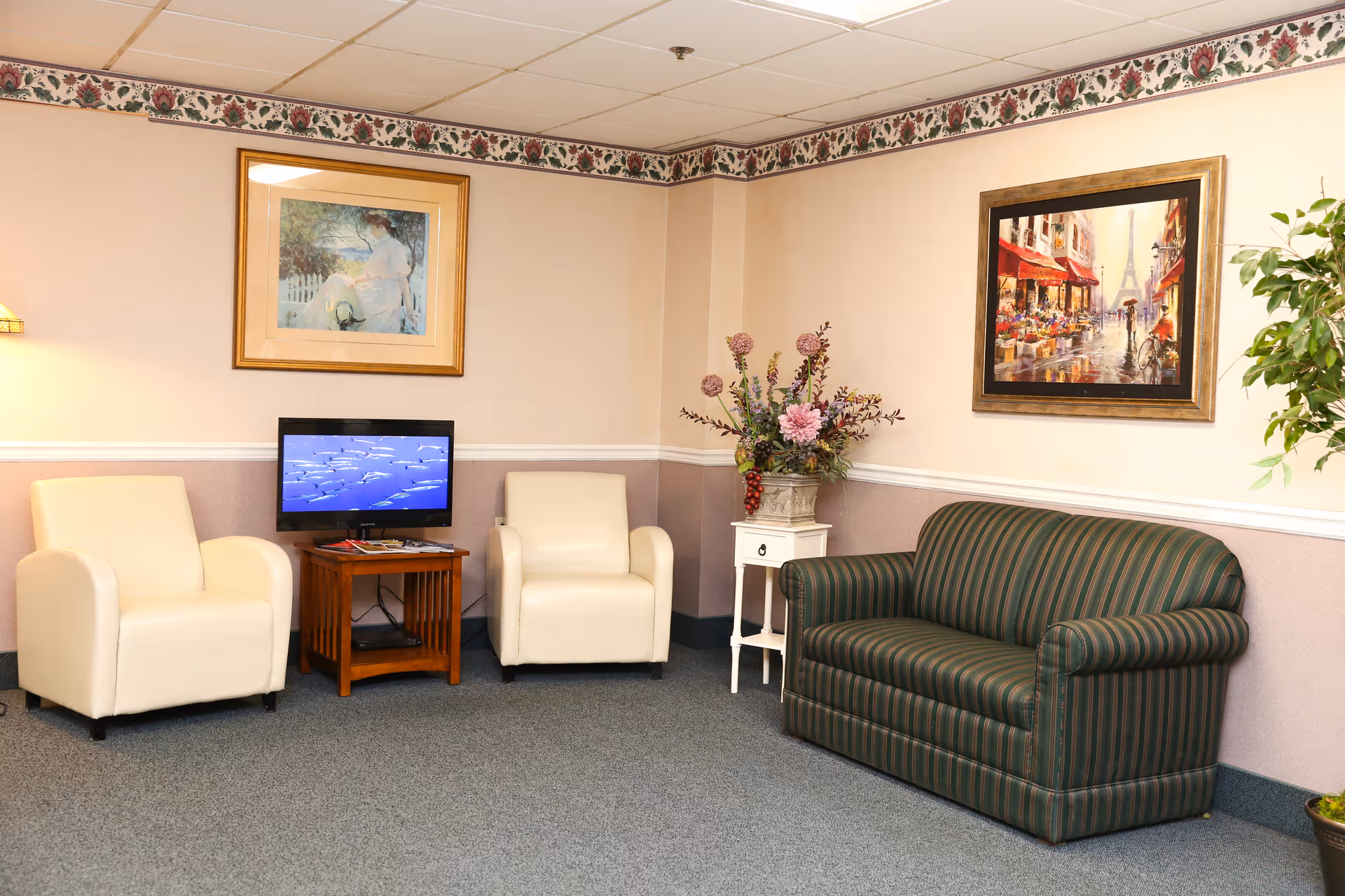 A cozy living room area with two cream-colored armchairs and a green striped sofa. A small wooden table with a TV displaying an underwater scene is placed between the armchairs. The walls are decorated with two framed paintings and a floral wallpaper border near the ceiling. A white side table with a floral arrangement and a potted plant add to the decor.