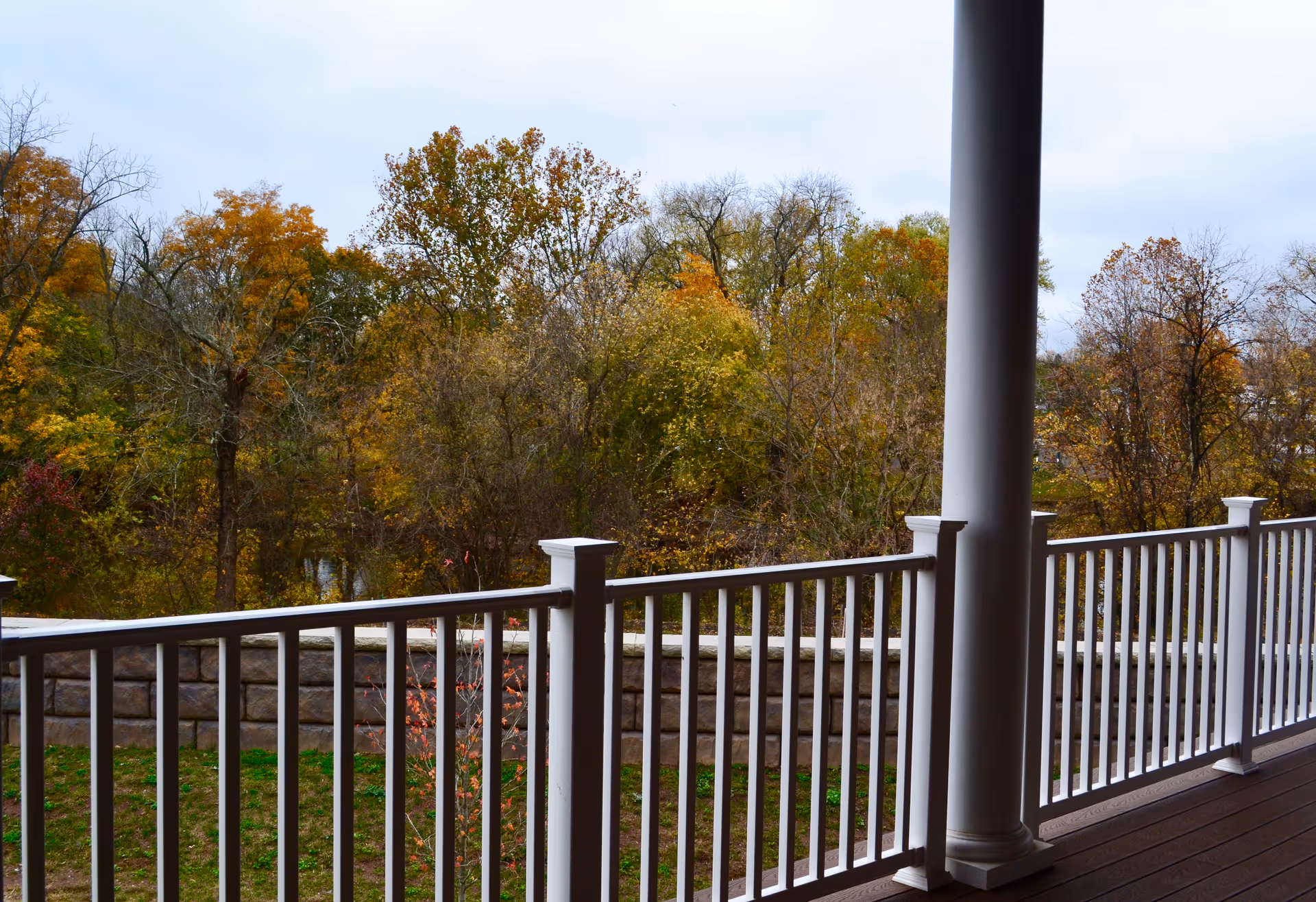 View from a porch with white railing and a large white column overlooking a grassy area and a stone retaining wall, with trees showing autumn foliage in the background under a cloudy sky.