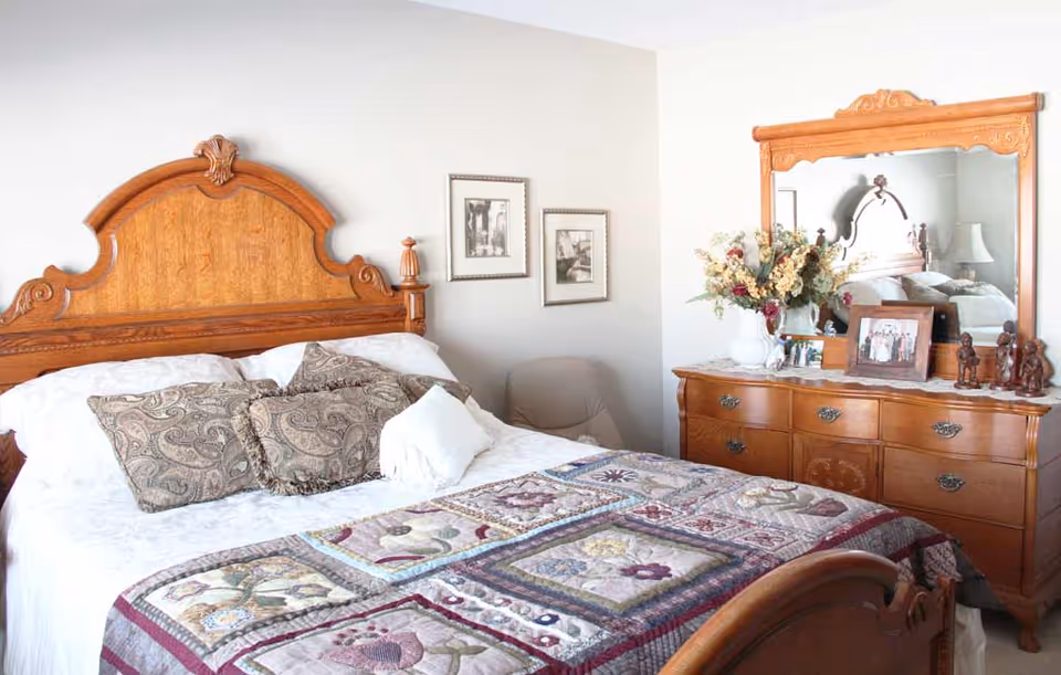 Sunlit bedroom with an ornate wooden bed and matching dresser topped with a mirror, flowers, and framed photos.