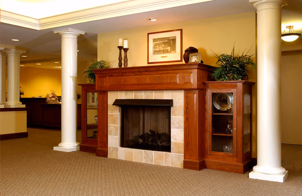 Interior view of a senior living facility featuring a wooden fireplace with a tiled surround, flanked by two wooden cabinets with glass doors displaying decorative items. The room has beige walls, carpeted flooring, white columns, and soft lighting, creating a warm and inviting atmosphere.