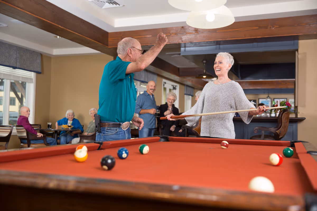 Two elderly people playing pool in a common room with a red pool table. The man in a teal shirt is raising his arm in celebration while the woman in a gray sweater holds a pool cue and smiles. In the background, several other elderly people are seated and standing, socializing in a well-lit room with wooden beams and large windows.