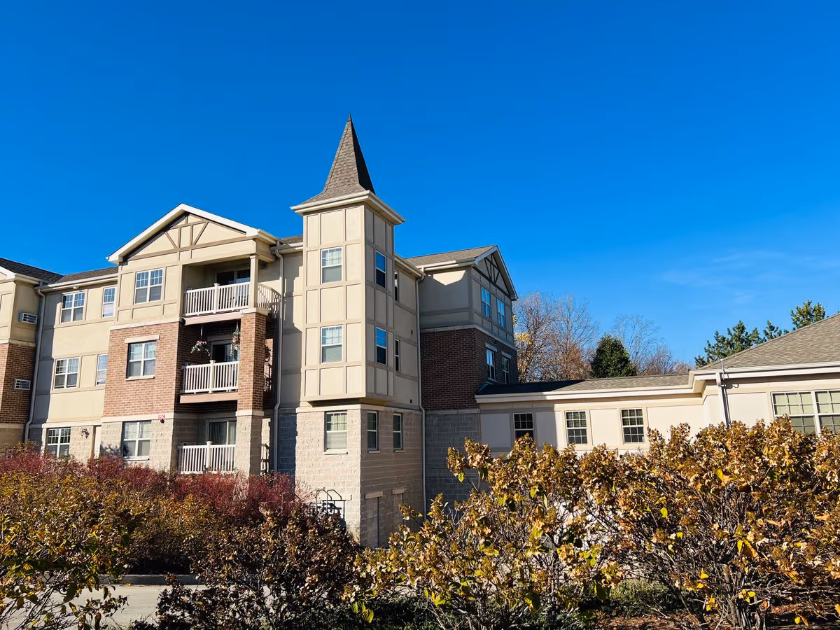 Exterior front of a multi-story senior living building with balconies, a turret-like tower, and shrubs in the foreground.