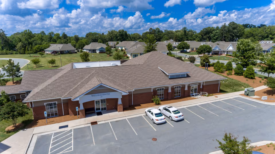 Aerial view of a brick wellness center labeled "WELLNESS CENTER" with two cars in the parking lot and surrounding residential buildings under a blue sky.