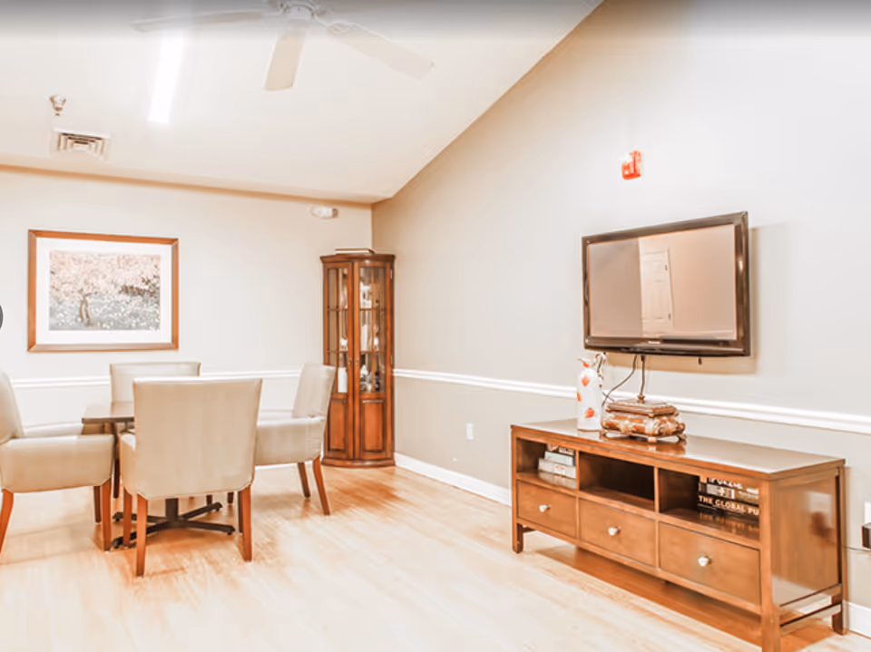 Bright common room with a dining table and chairs, a wall-mounted TV above a wooden console, and a glass display cabinet.
