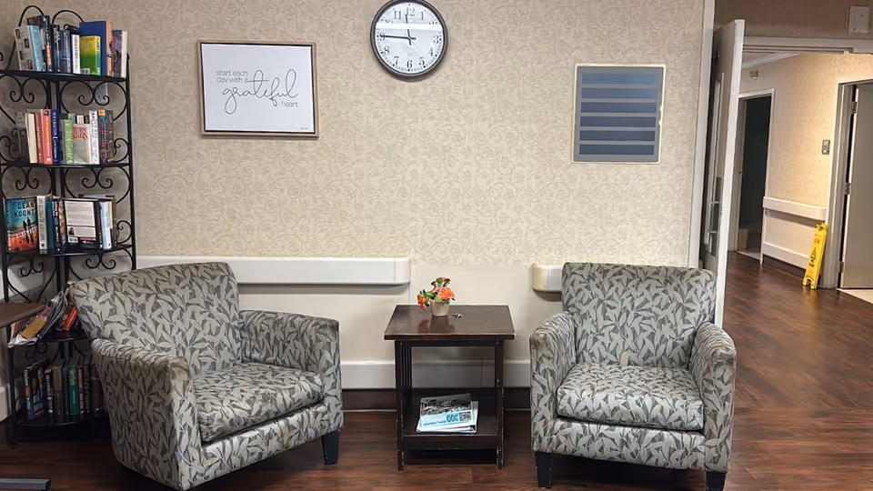Two patterned armchairs with a small table between them in a nursing-home sitting area, a bookshelf, wall clock and framed sign on the wall.