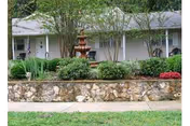Exterior view of a single-story building with a gray roof and white walls, surrounded by greenery including bushes, trees, and a stone wall in front. A multi-tiered water fountain is visible in the garden area.