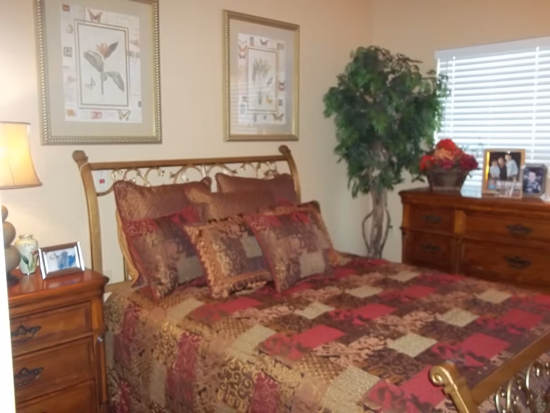 Well-decorated bedroom with a patterned bedspread, wooden nightstands and dresser, framed wall art, and a potted plant by a window.