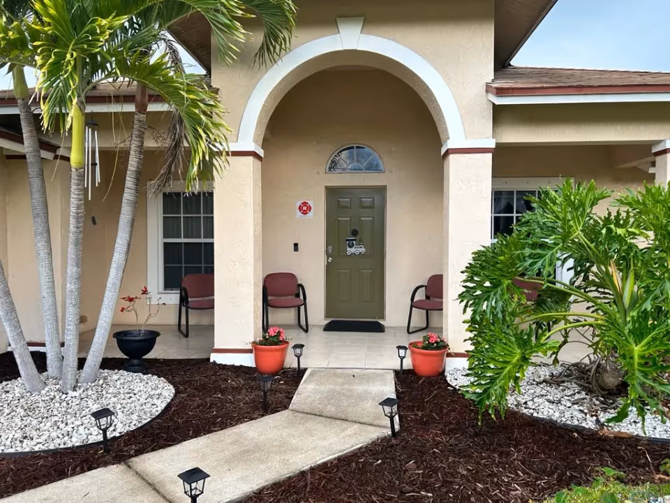 Beige facility entrance with an arched entryway, green door, two chairs on the porch, and tropical landscaping along a walkway.