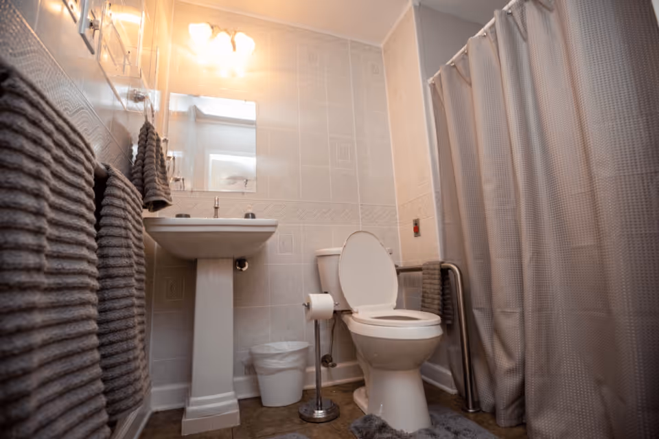 A clean bathroom with a white pedestal sink, a toilet with the lid open, a trash bin, and a shower area with a gray curtain. The walls are tiled in light colors, and there are gray towels hanging on the wall and a towel bar next to the toilet. A mirror and a light fixture with three bulbs are above the sink.