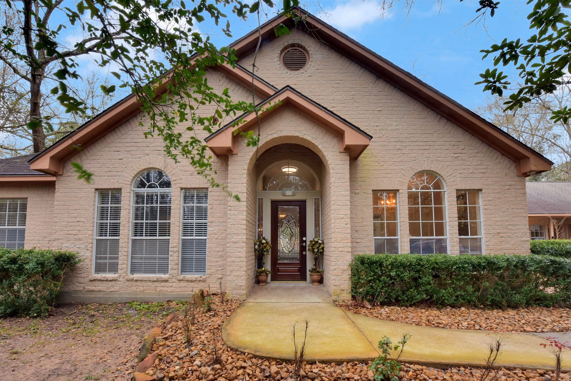 Front exterior view of a single-story brick building with a peaked roof, arched entryway, and decorative glass front door. The building has multiple large windows with white blinds and is surrounded by shrubs and a landscaped area with rocks and a concrete walkway leading to the entrance. Trees and a blue sky are visible in the background.