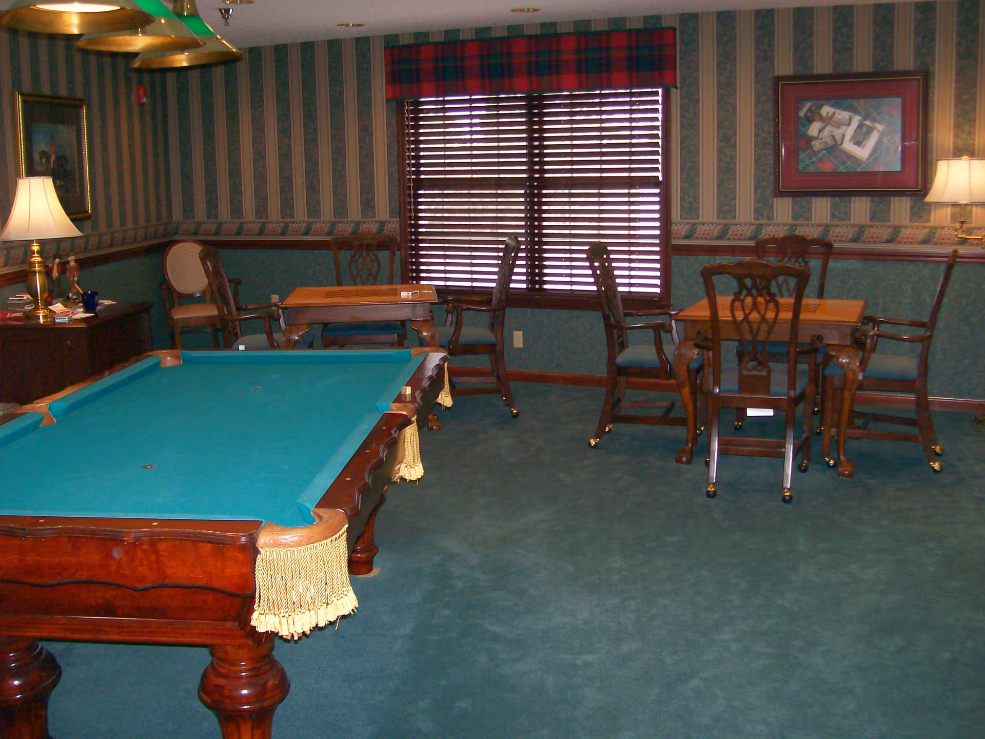 Pool table with green felt and several card tables and chairs in a traditional game room with striped wallpaper and window blinds.
