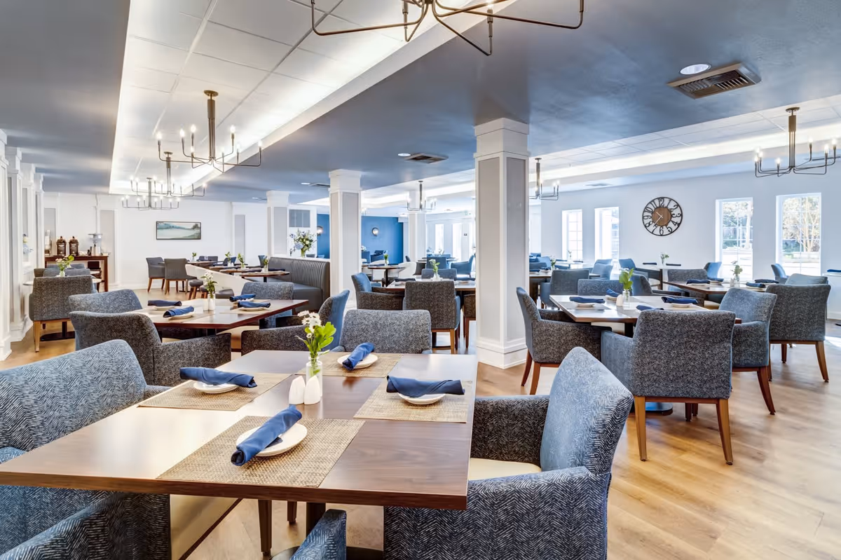 A spacious and well-lit dining room with multiple wooden tables set with blue napkins, white plates, and small flower vases. The room features comfortable upholstered chairs, light wood flooring, white walls, large windows allowing natural light, and modern chandeliers hanging from the ceiling.