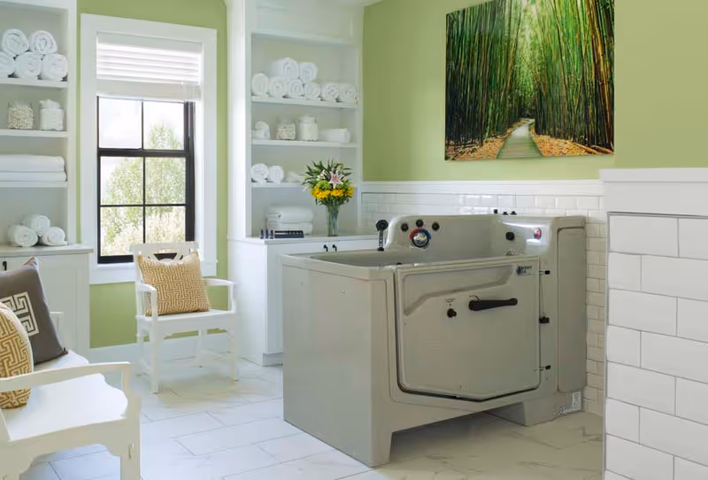 A bright and clean bathroom with a large walk-in bathtub, white shelves stocked with rolled towels and jars, a window letting in natural light, two white chairs with patterned cushions, and a large framed picture of a bamboo forest on a light green wall.