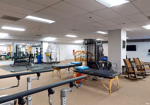 Interior view of a senior living facility's exercise and rehabilitation room featuring parallel bars for walking support, two padded therapy tables with blue cushions, exercise equipment, a large mirror on the wall, and a few wooden rocking chairs near a pillar.