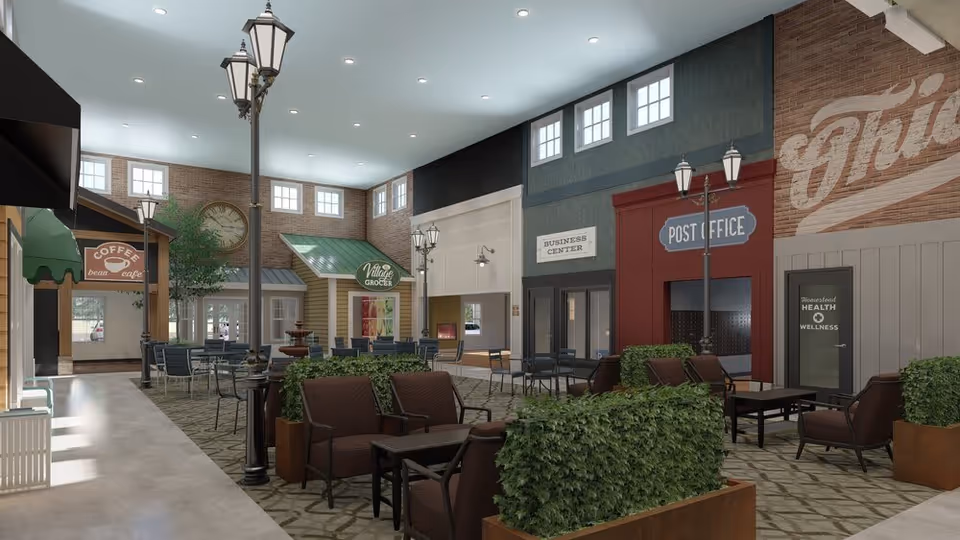 Spacious indoor common area styled like a small-town streetscape with seating, lampposts, and storefront facades including a post office, coffee shop, and grocer.