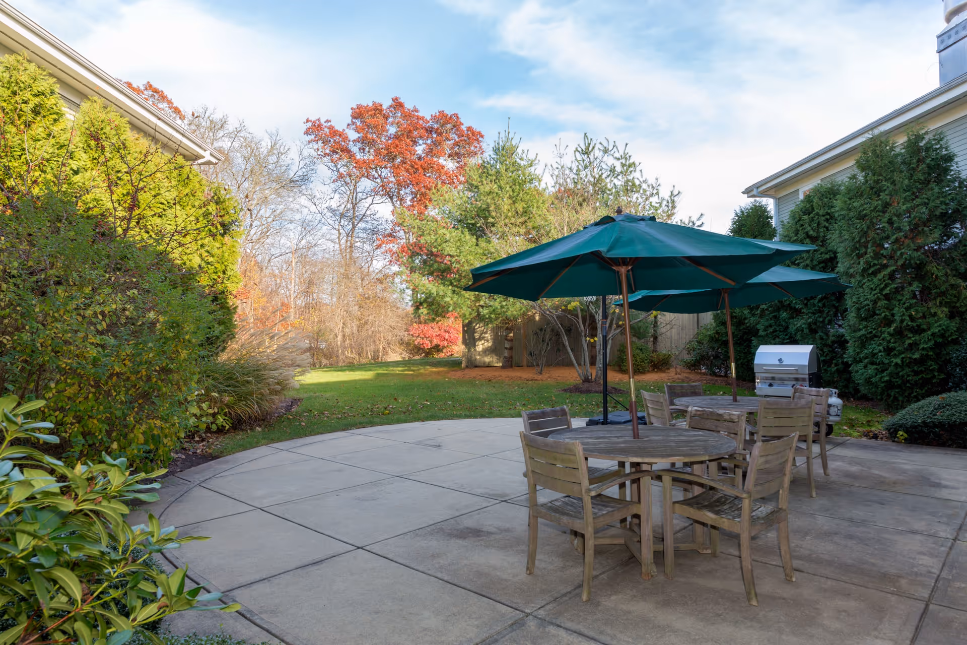 Outdoor patio area with round wooden tables and chairs under green umbrellas, surrounded by greenery and trees with autumn foliage. A barbecue grill is visible on the right side near the bushes.