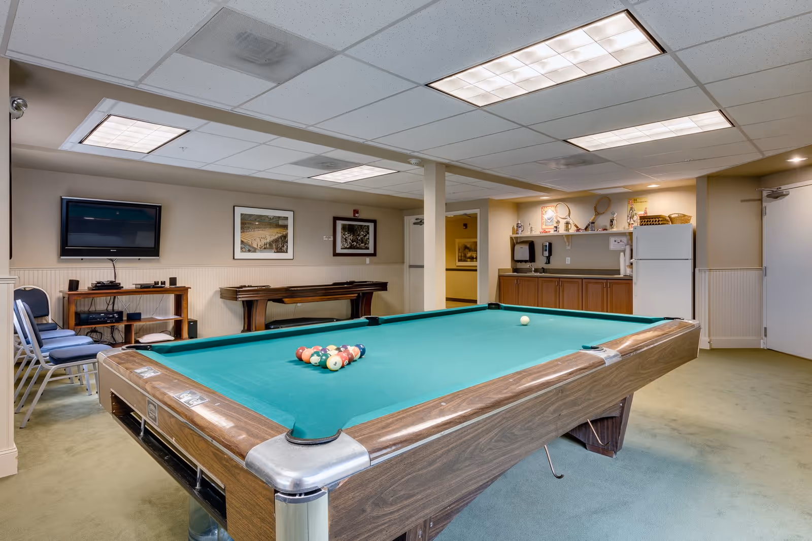 A recreational room with a pool table in the center, set up for a game with billiard balls arranged in a triangle. The room has a green carpet, beige walls, and a drop ceiling with fluorescent lighting. On the left side, there are stacked chairs and a wall-mounted flat-screen TV above a wooden shelf holding electronic devices. The back wall features framed pictures and a shuffleboard table. On the right side, there is a kitchenette area with wooden cabinets, a white refrigerator, and shelves holding various items including tennis rackets.