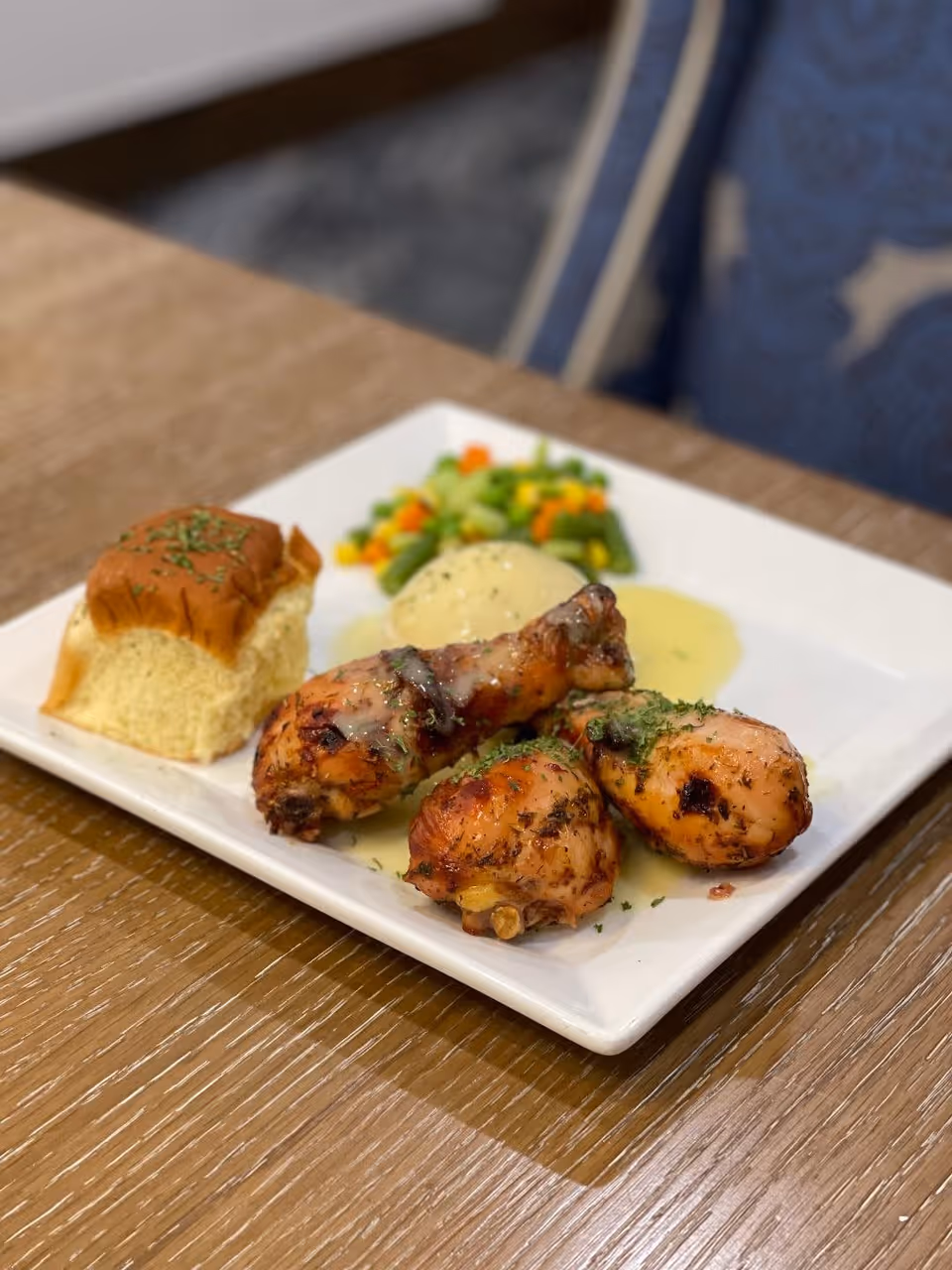 A plate of food on a wooden table featuring three grilled chicken drumsticks with herbs, a serving of mashed potatoes with gravy, mixed steamed vegetables, and a piece of dinner roll topped with herbs.