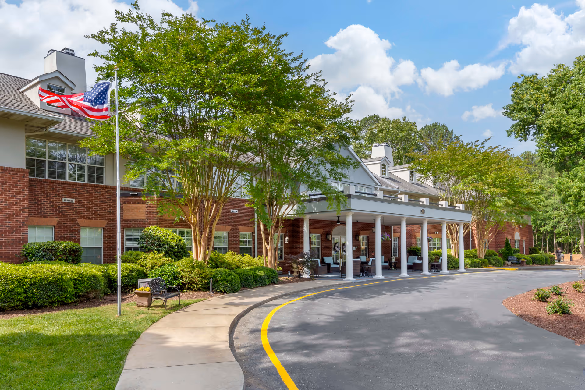 Exterior view of Brookdale MacArthur Park senior living facility showing a brick building with white columns at the entrance, a curved driveway with a yellow line, green trees and bushes, an American flag on a flagpole, and a partly cloudy sky.