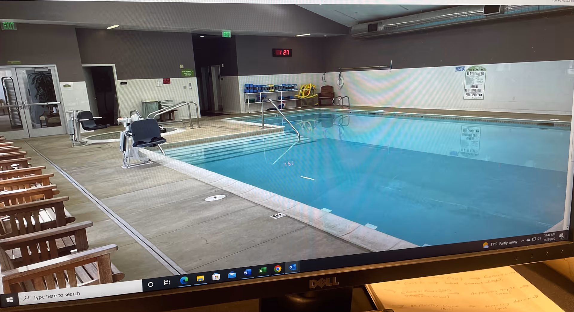 Indoor swimming pool area with clear blue water, pool lift chairs for accessibility, wooden chairs lined up along the left side, and a digital clock on the far wall showing 1:27. The pool area has safety signs on the walls and a door leading to other rooms.